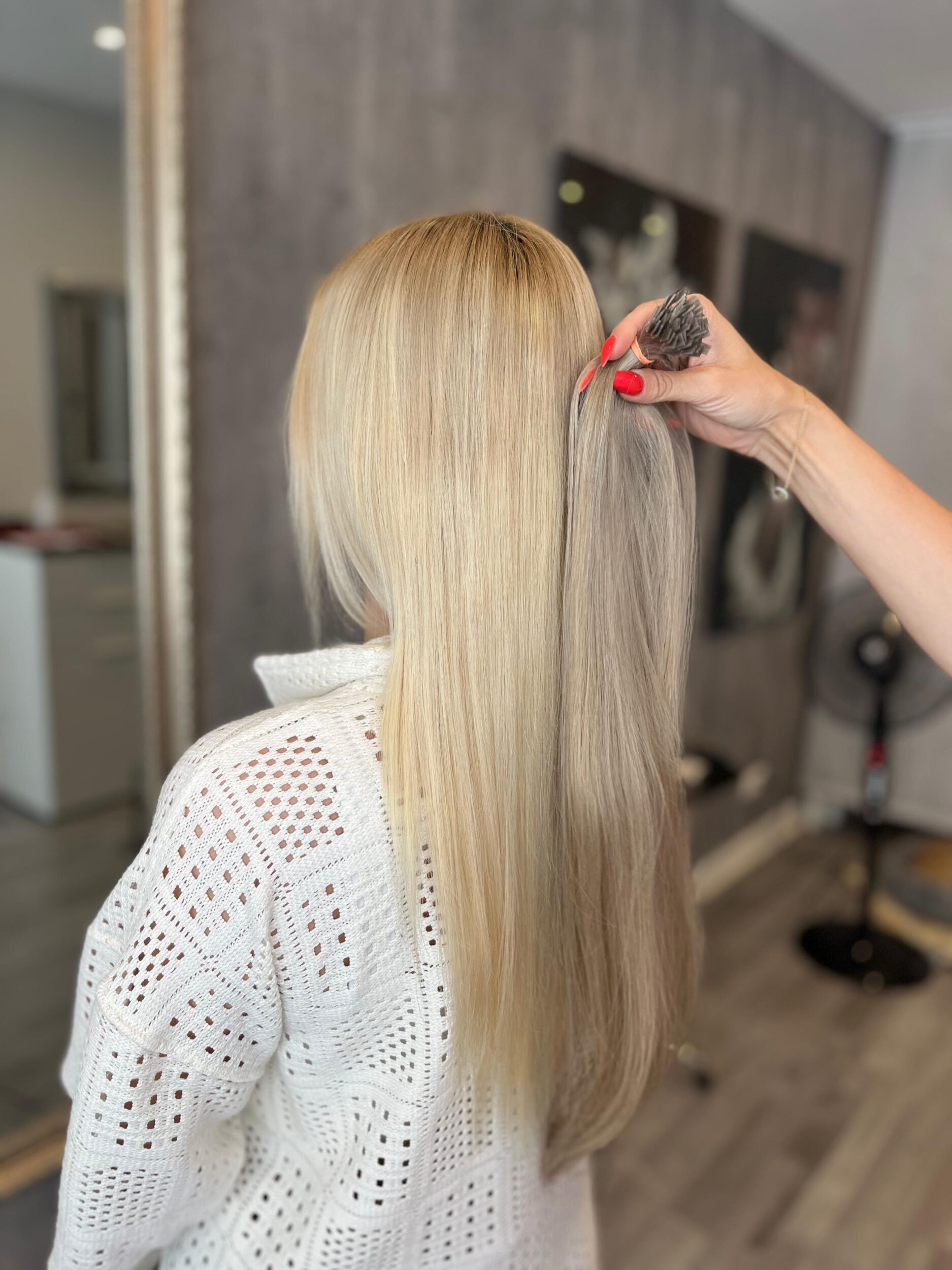 eine Blonde Frau vor dem Spiegel: an ire Haare wird eine Strähne Extensions gehalten