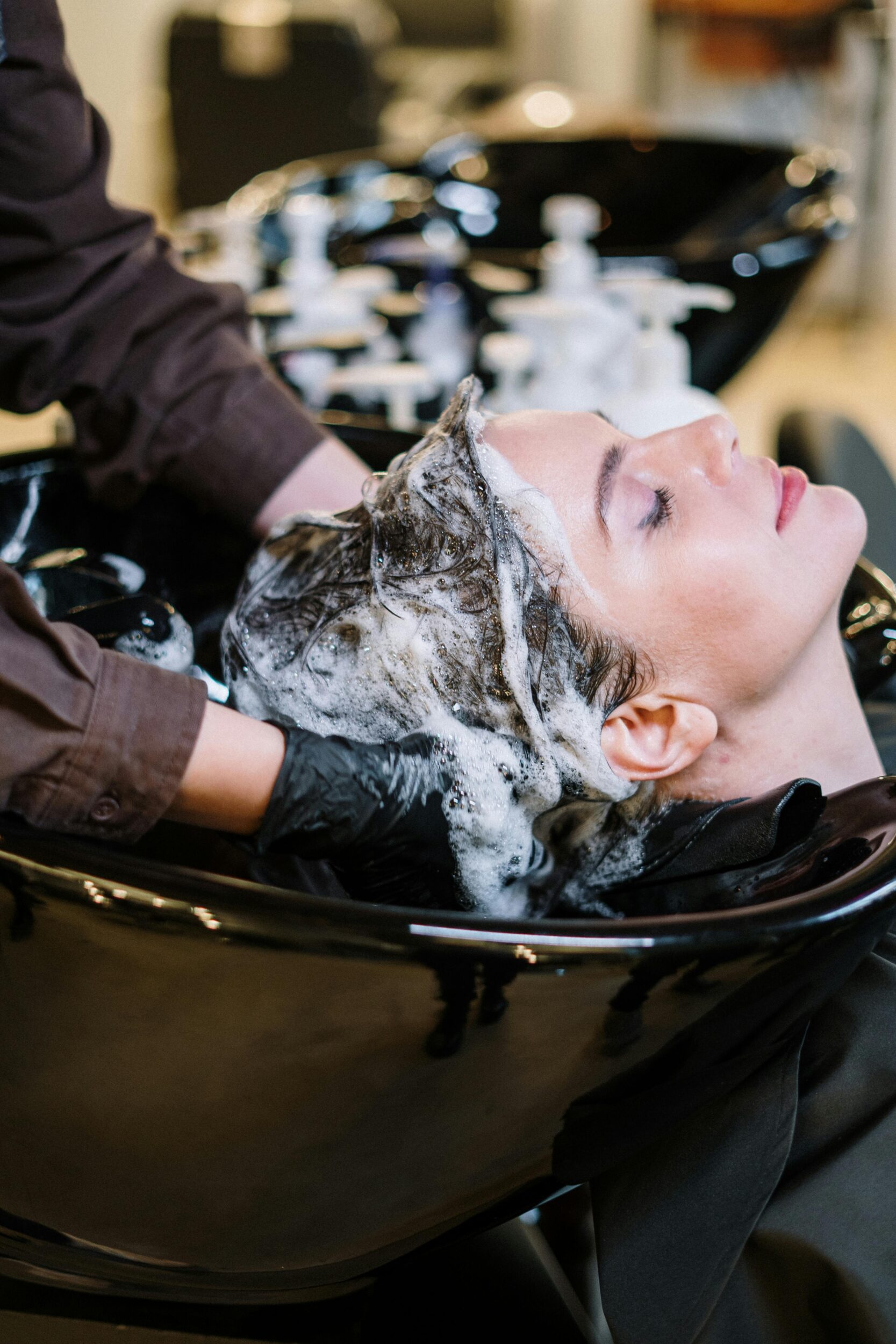 Person entspannt sich beim Haarewaschen im Friseursalon, während eine Friseurin mit schwarzen Handschuhen Shampoo einmassiert.